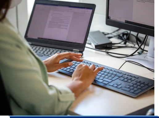 Eine Frau arbeitet an Ihrem Schreibtisch und bedient einen Computer. Zusätzlich hat sie einen Laptop neben sich stehen.
