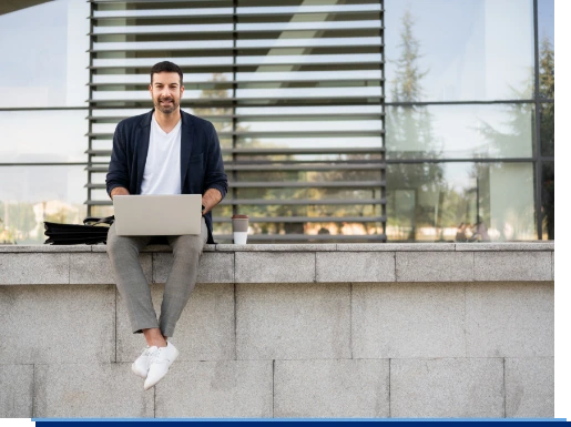 Ein Mitarbeiter sitzt im Freien auf einer Mauer und arbeitet entspannt über die Cloud an seinem Laptop.